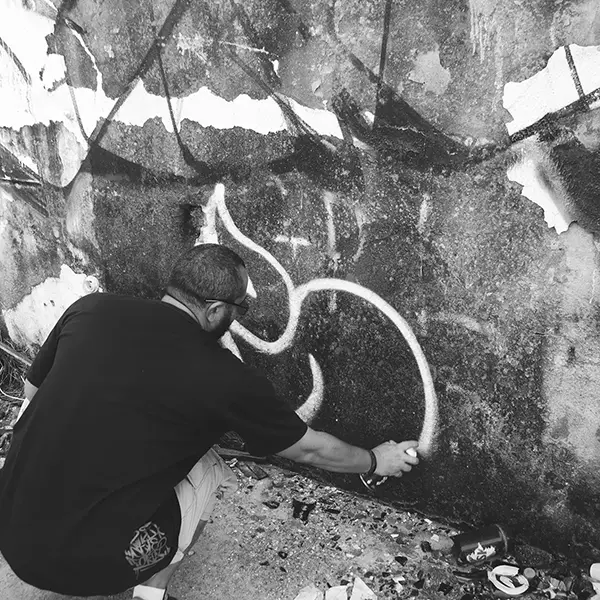 Frame 11 in grayscale from a video of a man squatting with a spray can in his hand while drawing a signature on the wall of an abandoned building