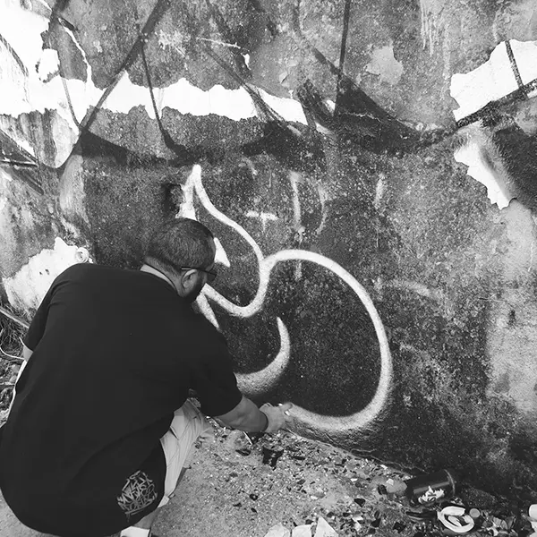 Frame 12 in grayscale from a video of a man squatting with a spray can in his hand while drawing a signature on the wall of an abandoned building