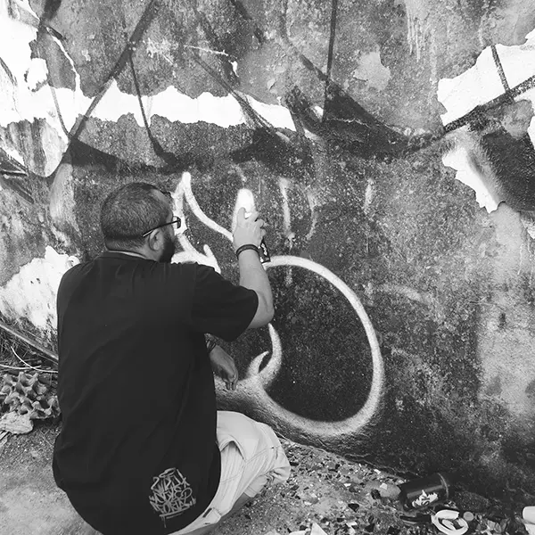 Frame 14 in grayscale from a video of a man squatting with a spray can in his hand while drawing a signature on the wall of an abandoned building