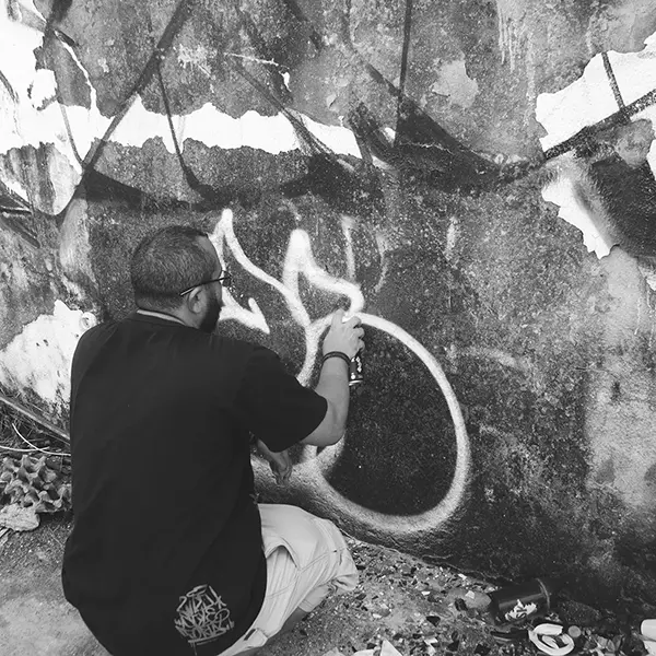 Frame 15 in grayscale from a video of a man squatting with a spray can in his hand while drawing a signature on the wall of an abandoned building