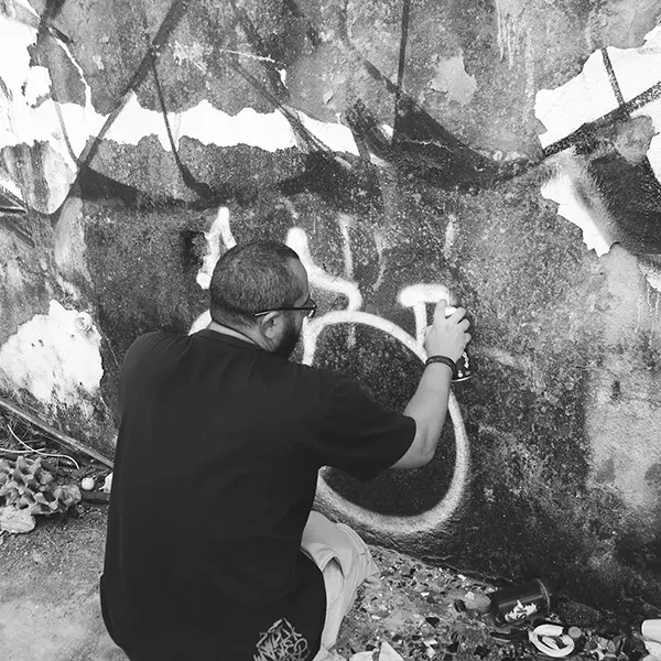 Frame 17 in grayscale from a video of a man squatting with a spray can in his hand while drawing a signature on the wall of an abandoned building