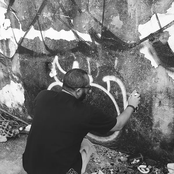 Frame 18 in grayscale from a video of a man squatting with a spray can in his hand while drawing a signature on the wall of an abandoned building
