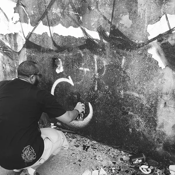 Frame 2 in grayscale from a video of a man squatting with a spray can in his hand while drawing a signature on the wall of an abandoned building