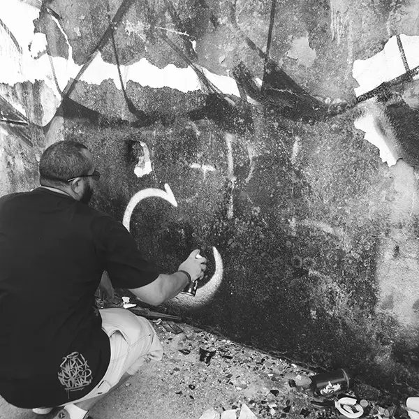 Frame 3 in grayscale from a video of a man squatting with a spray can in his hand while drawing a signature on the wall of an abandoned building