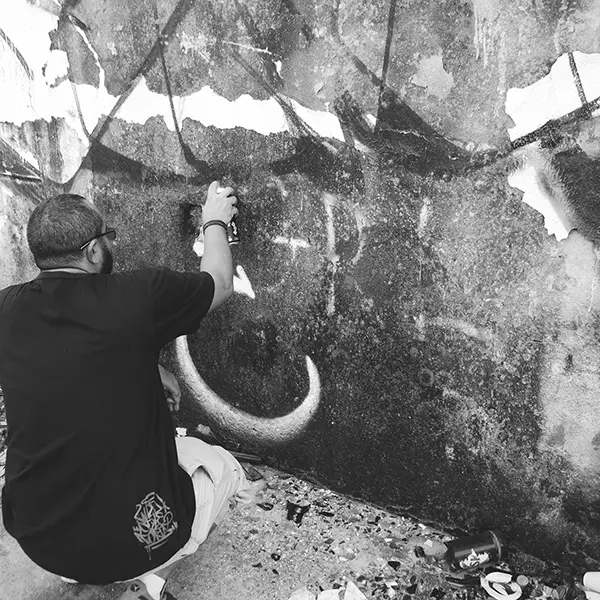 Frame 5 in grayscale from a video of a man squatting with a spray can in his hand while drawing a signature on the wall of an abandoned building