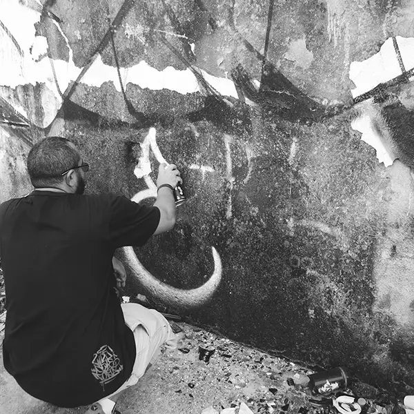 Frame 6 in grayscale from a video of a man squatting with a spray can in his hand while drawing a signature on the wall of an abandoned building