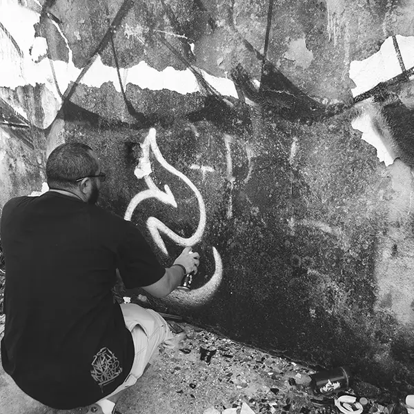 Frame 9 in grayscale from a video of a man squatting with a spray can in his hand while drawing a signature on the wall of an abandoned building